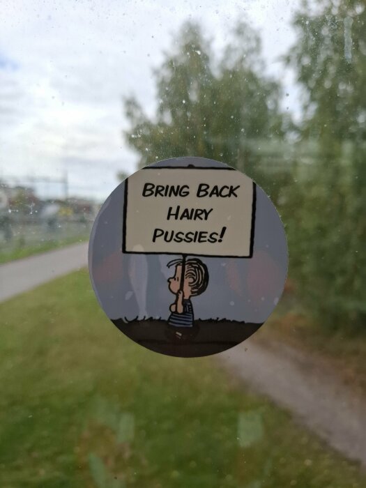 Dekal på fönster med en tecknad figur som håller upp en skylt med texten "Bring back hairy pussies!" i en utomhusmiljö.