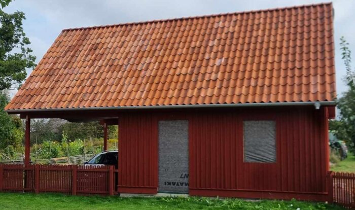 Kombinerat garage och carport under konstruktion med rött träpanel, tegelpannor på taket och tre öppna sidor, samt rektangulära fönsteröppningar.