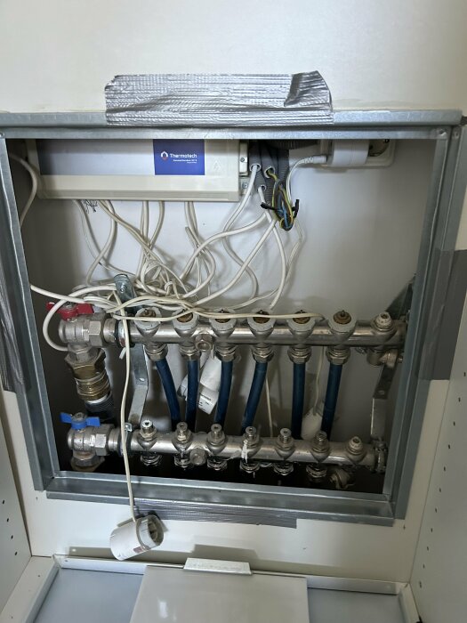 Distribution cabinet with blue water pipes and wiring, along with a control box labeled "Thermotech." A loose thermostat knob hangs below the pipes.