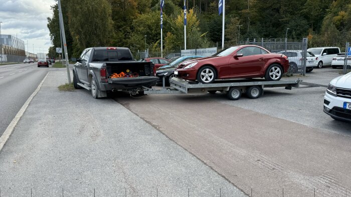 Grå pickup med öppet flak parkerad längs väg, bogserar bilsläp med röd sportbil. Bilar och skog i bakgrund.