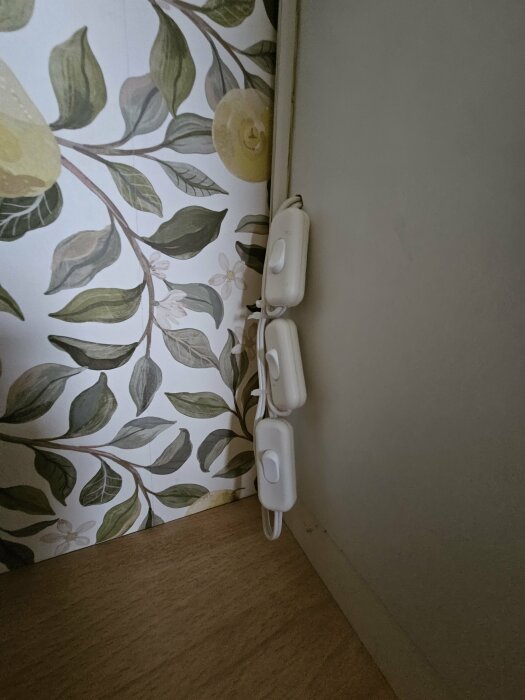 Tre vita strömbrytare kopplade till varsin kabel monterade på en vägg med blommig tapet.