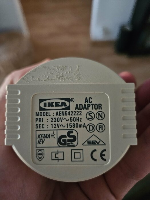 IKEA AC-adaptermodell AEN542222, visar primärt 230V~50Hz och sekundärt 12V~1580mA, hålls i handen mot suddig bakgrund.