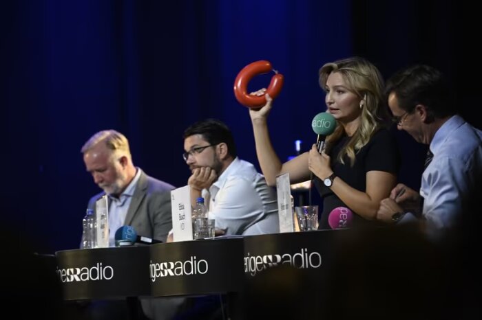 En paneldeltagare håller upp en röd korv medan hon pratar i en mikrofon under en debatt på Sveriges Radio. Fyra personer sitter vid ett bord med mikrofoner och vattenflaskor.