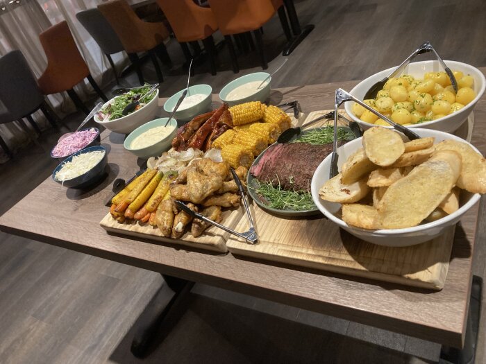 Buffébord med rotsaker, kycklingben, kött, majskolvar, potatis, sallad, vit och rosa sås samt vita och orangefärgade stolar i bakgrunden.