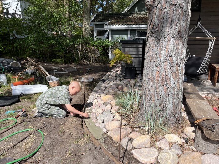 Barn som placerar en betongkant bredvid en gräsmatta. Stenar och ett snår syns i bakgrunden med en trädstam till höger i bilden.