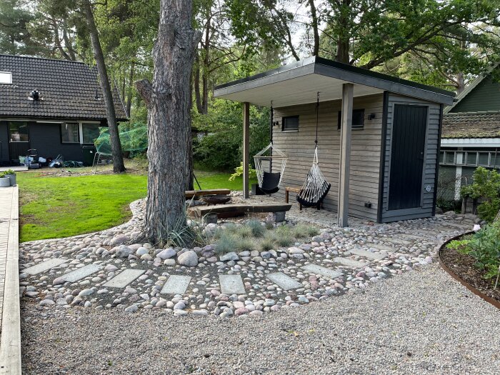 Grått träskjul med svart dörr och sittplatser under taket, omgiven av rundad stengång med plattor, gräs och träd i trädgård. En hammock hänger framför skjulet.