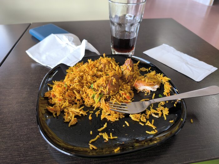 Svart tallrik med Chicken Biryani, bitar av kyckling och ris, på ett bord med en dricksglas och servetter bredvid.