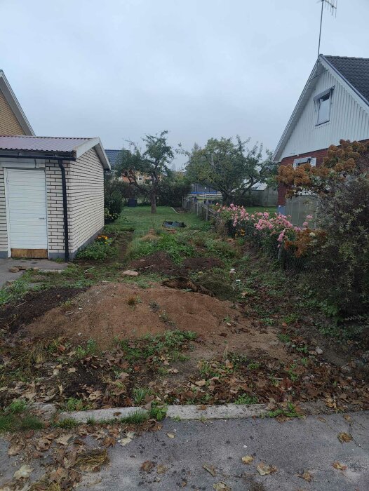 Trädgårdsområde med gräsmatta, buskar och jordhögar framför ett garage och ett hus, i samband med garageproblemen som beskrivs.