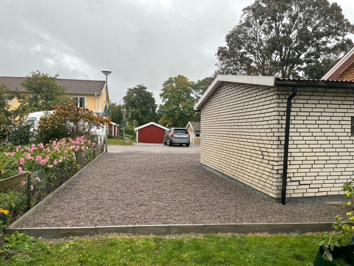 Ny grusad uppfart framför ett garage med sprucken fasad. Blommor vid staketet och hus i bakgrunden. Grå himmel ovanför.