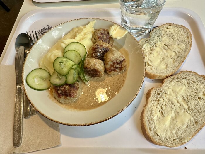 Köttbullar med gräddsås och potatismos, garnerade med skivad gurka, serverade med smörat bröd och ett glas vatten på en bricka.