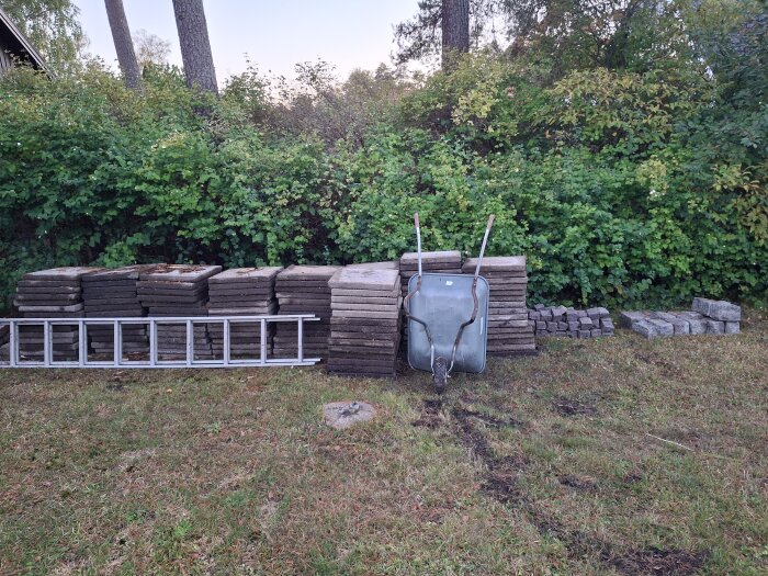 Staplar av sjöstensplattor, gatsten och en skottkärra står framför en grön buske, med en stege liggande på marken intill.