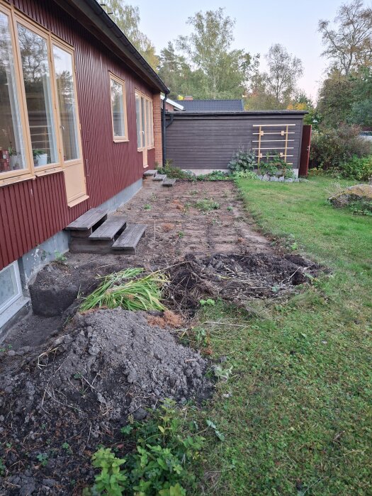 Grävd yta bredvid rött hus med träväggar, förberett för dränering. Jordhögar och vissna växter syns, med trappsteg av trä mot huset.