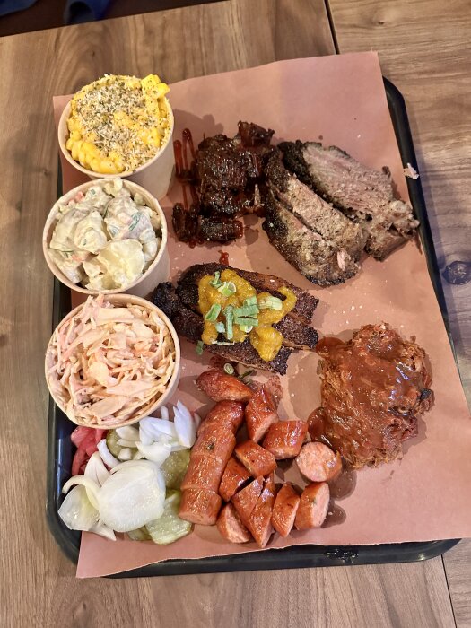 En serveringsbricka med mac and cheese, potatissallad, coleslaw, grillad korv, och kött inklusive pulled pork och burnt ends, för tre personer.