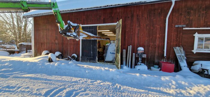 Grön teleskoplastare vid röd lada i snöigt landskap, med en stor säck lastad genom ladans dörr.