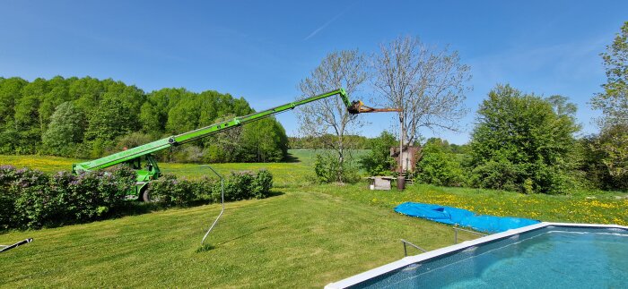 Grön teleskoplastare sträcker sig över gräsmatta mot träd, sommarklädd omgivning med pool, träd och blå himmel.