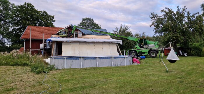 Grön teleskoplastare vid byggarbetsplats, använd för att montera tak på rött hus med grönskande träd i bakgrunden.