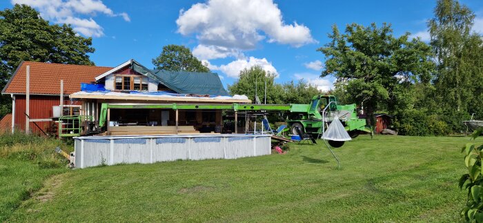 En grön teleskoplastare står bredvid ett hus med renovering pågående, omgiven av gräs och träd under en blå himmel.