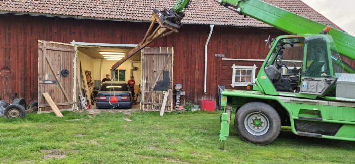 Grön teleskoplastare framför rött trähus, används för att lyfta en bil i ett garage med öppna dörrar. Två personer står nära bilen.