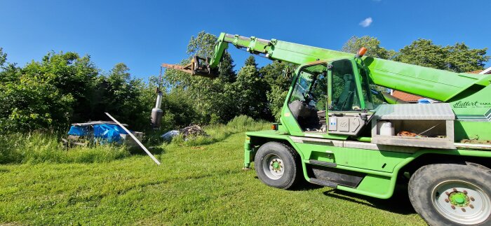 Grön teleskoplastare i trädgård lyfter objekt framför buskar med blå himmel i bakgrunden.