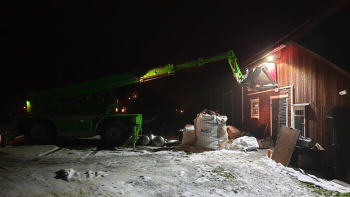 En grön teleskoplastare används för att lyfta material till en träbyggnad på vintern, omgiven av snö och byggnadsmaterial.