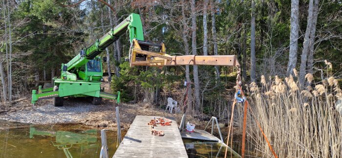 Grön teleskoplastare vid sjön, med lyftarm utsträckt över brygga och gräs vid strandkanten i skogsmiljö.