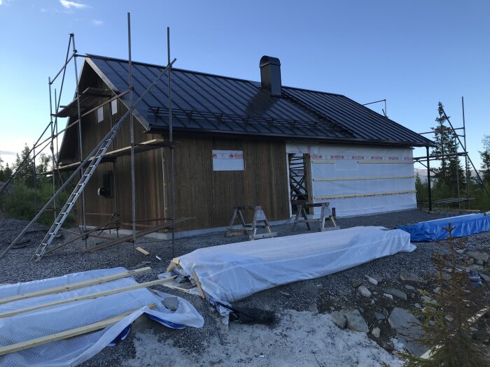 Trähus under konstruktion med svart tak, ställningar och material på marken. Isolerad skorsten och plåttakhylsa synliga.