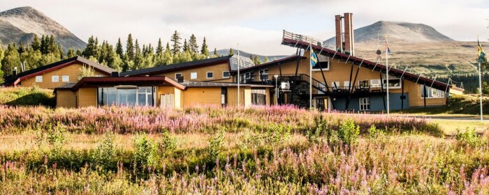 Hotell Borgafjäll i fjällandskap, med skidbacke på taket och omgiven av blommande ängar och skog i bakgrunden.