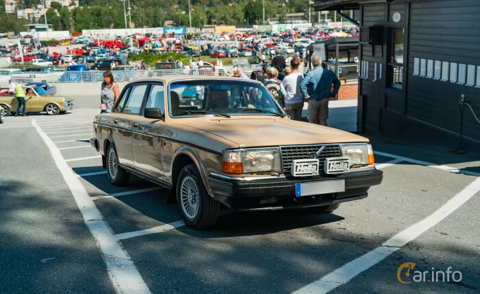 Volvo 240 med rektangulära Hella-extraljus monterade framtill, parkerad på en bilträff med folk och bilar i bakgrunden.