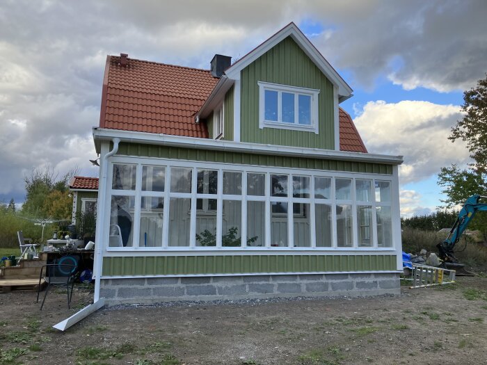 Grönt hus med ryggåstak och nybyggt växthus på gjuten betongsockel, omgiven av trädgårdsutrustning och grävmaskin i bakgrunden.