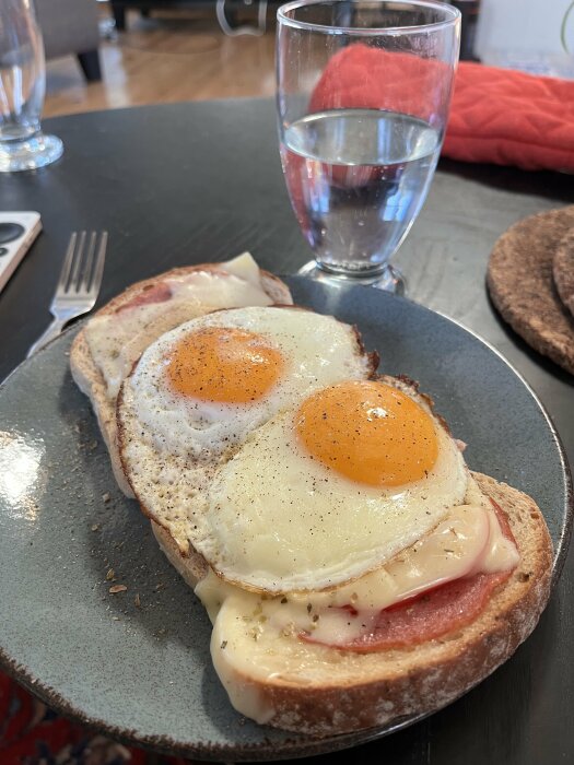 Smörgås med medvurst, smält ost, stekt ägg, tomat och paprika, toppad med peppar och oregano, serverad på en tallrik med ett glas vatten.