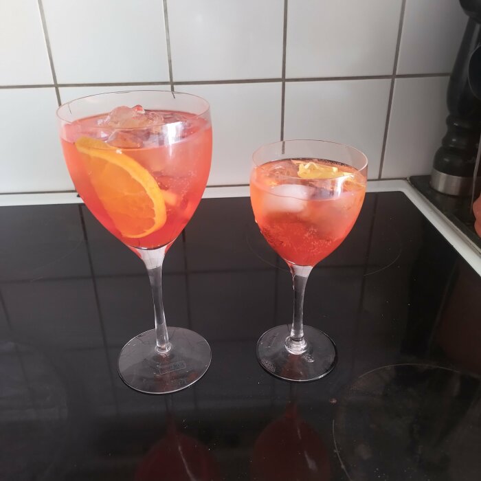 Två glas Aperol spritz, ett stort och ett litet, med apelsinskivor och isbitar på en spishäll.