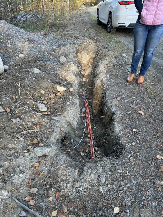 En person står bredvid ett dike fyllt med röd kanalisation för optisk fiber vid en skogsväg. En vit bil syns i bakgrunden.