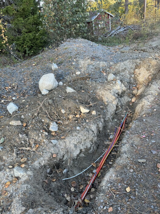 Dike för fiberkanalisation på en tomt med synliga kablar i marken, omgiven av skog och grus.