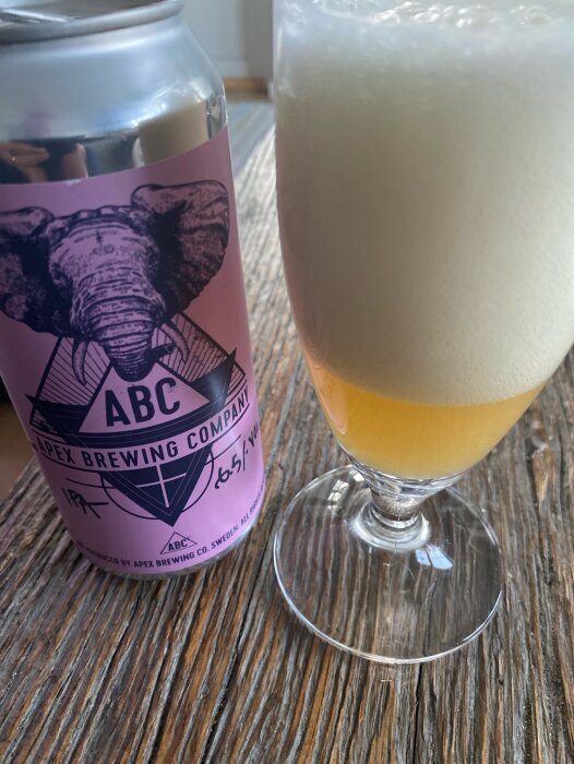 En burk Apex IPA med elefantlogo bredvid ett glas fyllt med skummande öl på ett träbord.