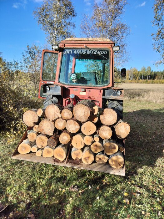 Röd traktor med vedlast på fält, höstlandskap i bakgrunden.