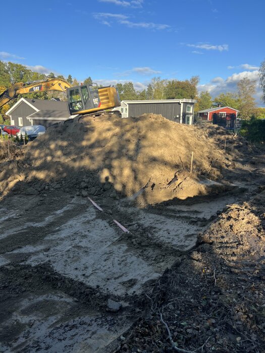 Schaktmassor av jord och sten ligger i en hög på en tomt, med en grävmaskin ovanpå och hus i bakgrunden under klar himmel.