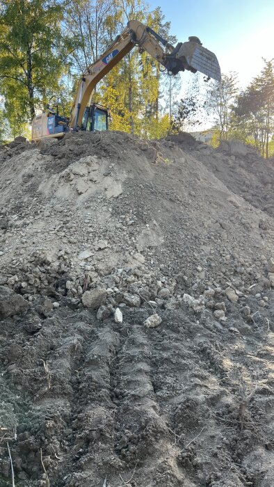 Grävskopa placerar sten- och jordmassor i hög i en skogsmiljö, potentiellt för användning som fyllnadsmaterial vid byggprojekt.