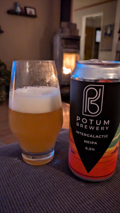 Ölglas med skumfylld dryck bredvid en burk Potum Brewery Intergalactic NEIPA 6,5%, bakgrund med brasa och inomhusmiljö.