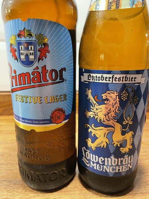 Två ölflaskor, en med etiketten "Primátor Festive Lager" och en "Löwenbräu Oktoberfestbier", står på ett träbord.