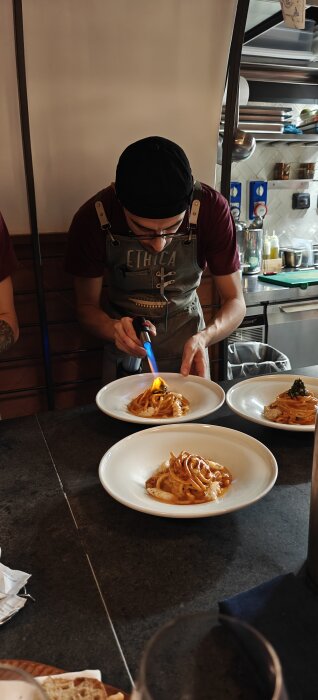 Kock på restaurang som bränner ytan på pasta med en brännare, servering förbereds i köket.