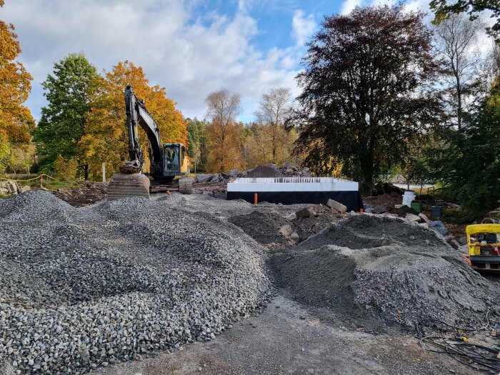 Återfyllnad av källare pågår, grushög i förgrund, grävmaskin i bakgrund och nyligen gjuten garageplatta vid byggarbetsplats.