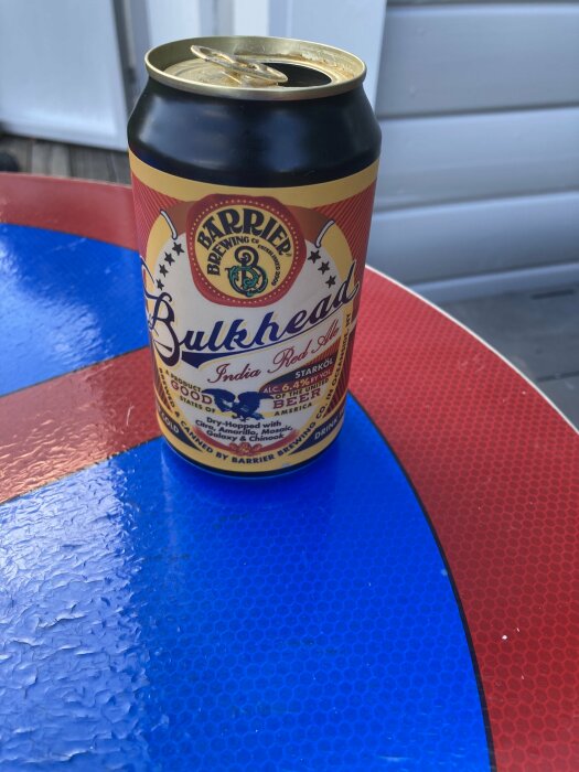 En ölburk med etiketten "Bulkhead India Red Ale" står på ett färgglatt bord.