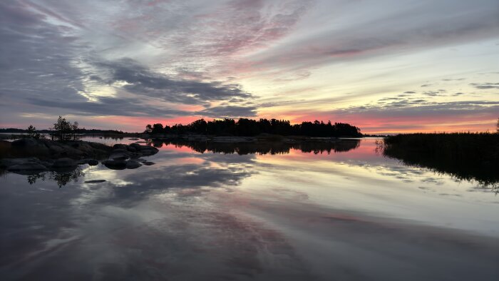 Sjön reflekterar en höstsolnedgång med rosa och orange färger över en skog och klippor, vilket skapar en stämningsfull naturvy.