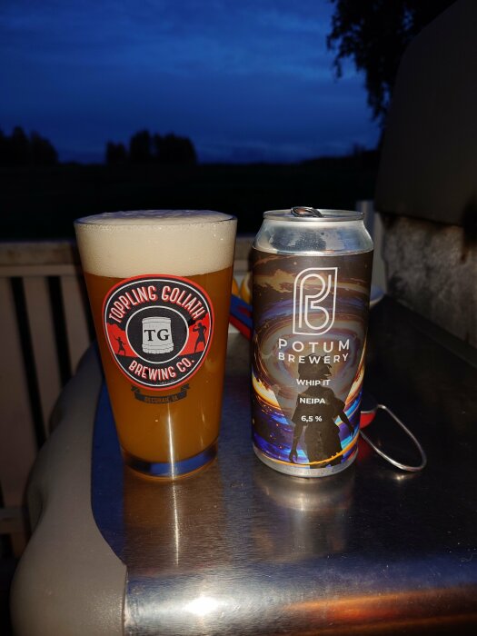 Glas med öl från Toppling Goliath-bryggeri bredvid en burk Potum Brewery NEIPA på en bänkyta mot kvällshimlen i bakgrunden.