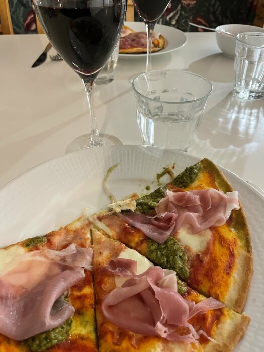 Pizza med mozzarella, pesto och skinka serverad på tallrik, omgiven av två glas rött vin och ett glas vatten på ett vitt bord.