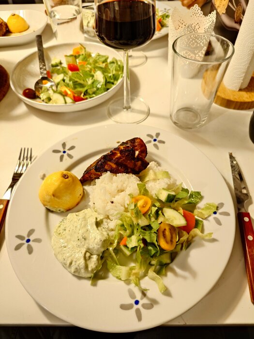 Tallrik med grillad kyckling, ris, basilikasås, sallad och ett glas rödvin på ett dukat bord.