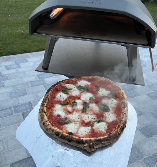 Pizza framför en pizzaugn på en stenlagd uteplats, toppad med mozzarella och basilika. Pizzaugnen är ny och användaren är mycket nöjd.