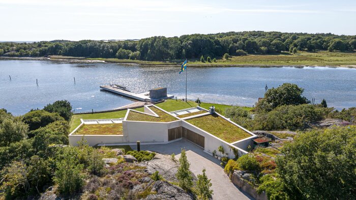 Modern villa med sodatak vid vattnet, svensk flagga, brygga och grönskande omgivningar i naturskön miljö.