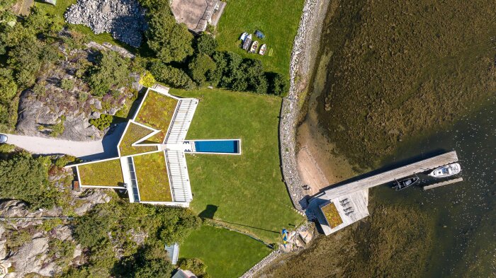 Flygfoto av arkitektoniskt hus med grönt tak, stor trädgård, pool och brygga med båtar vid sjön. Omringat av natur och sten.