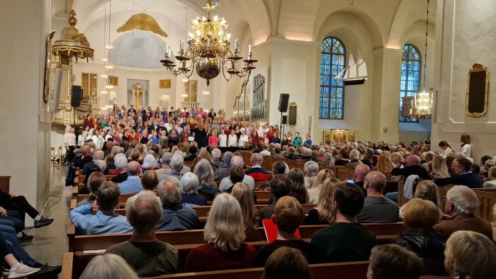 En stor kör framför en publik i en kyrka med en ljuskrona och orgelpipor i bakgrunden.